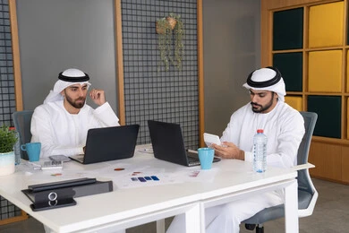 Integrating work with technology, the development of Emirati companies, following up on business projects, two Emirati Gulf Arab men wearing kandura and ghutra completing work tasks via computer, expressions and gestures of concentration, working on a plan to achieve the company's goals