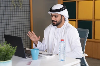 مكالمات فيديو وبث مباشر, التلويح باليد والسلام عن بعد، رجل عربي خليجي اماراتي يرتدي الغترة والكندورة يقوم باستخدام جهاز الحاسوب المحمول, عقد صفقات عمل خارجية، اجتماعات عمل أونلاين، دمج العمل بالتقنية والتكنولوجيا، شركات الامارات العربية المتحدة