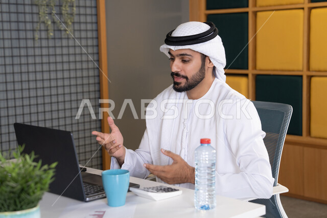 عقد صفقات عمل خارجية، اجتماعات عمل أونلاين، دمج العمل بالتقنية والتكنولوجيا، شركات الامارات العربية المتحدة، بث مباشر ومكالمات فيديو، التلويح باليد والسلام عن بعد، رجل عربي خليجي اماراتي يرتدي الغترة والكندورة يقوم باستخدام جهاز الحاسوب المحمول