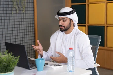 عقد صفقات عمل خارجية، اجتماعات عمل أونلاين، دمج العمل بالتقنية والتكنولوجيا، شركات الامارات العربية المتحدة، بث مباشر ومكالمات فيديو، التلويح باليد والسلام عن بعد، رجل عربي خليجي اماراتي يرتدي الغترة والكندورة يقوم باستخدام جهاز الحاسوب المحمول