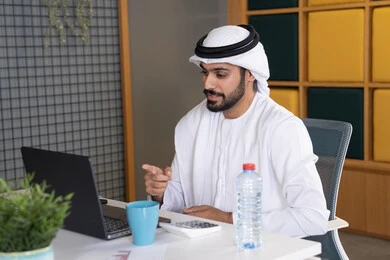 اجتماعات عمل أونلاين، دمج العمل بالتقنية والتكنولوجيا، شركات الامارات العربية المتحدة، بث مباشر ومكالمات فيديو، التلويح باليد والسلام عن بعد، رجل عربي خليجي اماراتي يرتدي الغترة والكندورة يقوم باستخدام جهاز الحاسوب المحمول، عقد صفقات عمل خارجية