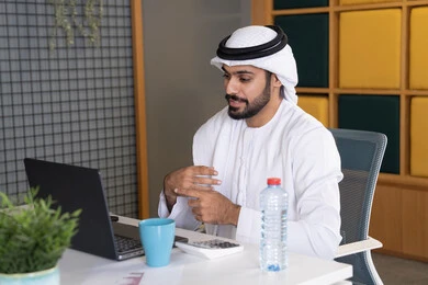 دمج العمل بالتقنية والتكنولوجيا، شركات الامارات العربية المتحدة، بث مباشر ومكالمات فيديو، التلويح باليد والسلام عن بعد، رجل عربي خليجي اماراتي يرتدي الغترة والكندورة يقوم باستخدام جهاز الحاسوب المحمول، عقد صفقات عمل خارجية، اجتماعات عمل أونلاين