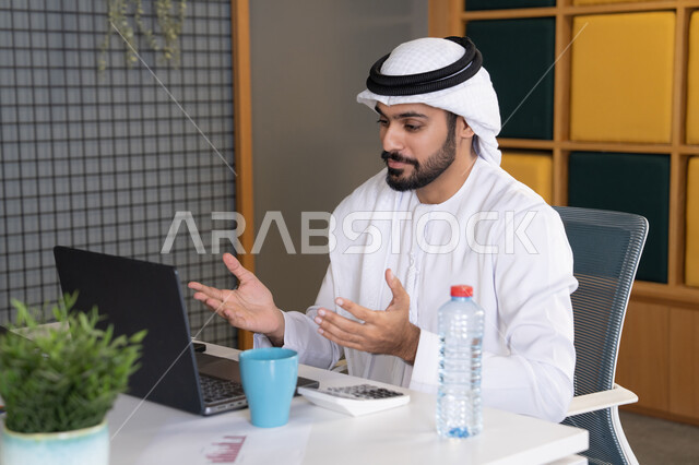عقد صفقات عمل خارجية، اجتماعات عمل أونلاين، دمج العمل بالتقنية والتكنولوجيا، شركات الامارات العربية المتحدة، بث مباشر ومكالمات فيديو، التلويح باليد والسلام عن بعد، رجل عربي خليجي اماراتي يرتدي الغترة والكندورة يقوم باستخدام جهاز الحاسوب المحمول