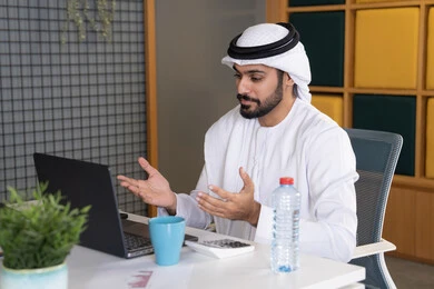 عقد صفقات عمل خارجية، اجتماعات عمل أونلاين، دمج العمل بالتقنية والتكنولوجيا، شركات الامارات العربية المتحدة، بث مباشر ومكالمات فيديو، التلويح باليد والسلام عن بعد، رجل عربي خليجي اماراتي يرتدي الغترة والكندورة يقوم باستخدام جهاز الحاسوب المحمول