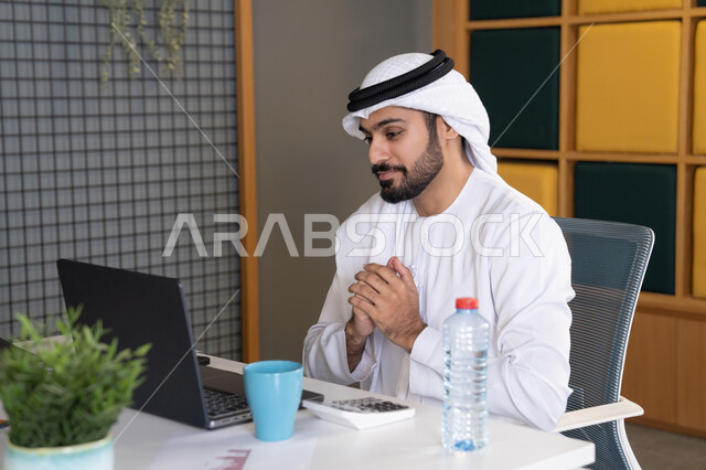 اجتماعات عمل أونلاين، دمج العمل بالتقنية والتكنولوجيا، شركات الامارات العربية المتحدة، بث مباشر ومكالمات فيديو، التلويح باليد والسلام عن بعد، رجل عربي خليجي اماراتي يرتدي الغترة والكندورة يقوم باستخدام جهاز الحاسوب المحمول، عقد صفقات عمل خارجية