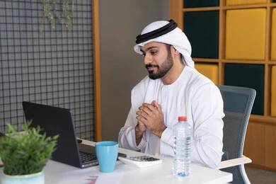 اجتماعات عمل أونلاين، دمج العمل بالتقنية والتكنولوجيا، شركات الامارات العربية المتحدة، بث مباشر ومكالمات فيديو، التلويح باليد والسلام عن بعد، رجل عربي خليجي اماراتي يرتدي الغترة والكندورة يقوم باستخدام جهاز الحاسوب المحمول، عقد صفقات عمل خارجية