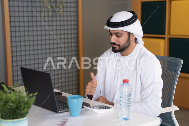 مكالمات فيديو وبث مباشر, التلويح باليد والسلام عن بعد، رجل عربي خليجي اماراتي يرتدي الغترة والكندورة يقوم باستخدام جهاز الحاسوب المحمول, عقد صفقات عمل خارجية، اجتماعات عمل أونلاين، دمج العمل بالتقنية والتكنولوجيا، شركات الامارات العربية المتحدة