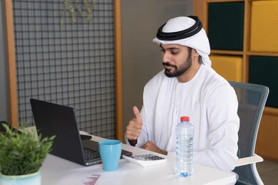 مكالمات فيديو وبث مباشر, التلويح باليد والسلام عن بعد، رجل عربي خليجي اماراتي يرتدي الغترة والكندورة يقوم باستخدام جهاز الحاسوب المحمول, عقد صفقات عمل خارجية، اجتماعات عمل أونلاين، دمج العمل بالتقنية والتكنولوجيا، شركات الامارات العربية المتحدة
