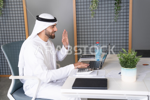 التلويح باليد والسلام عن بعد، رجل عربي خليجي اماراتي يرتدي الغترة والكندورة يقوم باستخدام جهاز الحاسوب المحمول, عقد صفقات عمل خارجية، اجتماعات عمل أونلاين، دمج العمل بالتقنية والتكنولوجيا، شركات الامارات العربية المتحدة، بث مباشر ومكالمات فيديو