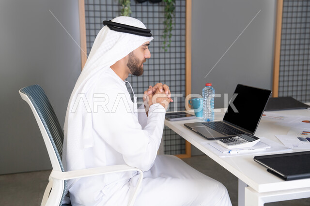 دمج العمل بالتقنية والتكنولوجيا، شركات الامارات العربية المتحدة، بث مباشر ومكالمات فيديو، التلويح باليد والسلام عن بعد، رجل عربي خليجي اماراتي يرتدي الغترة والكندورة يقوم باستخدام جهاز الحاسوب المحمول، عقد صفقات عمل خارجية، اجتماعات عمل أونلاين