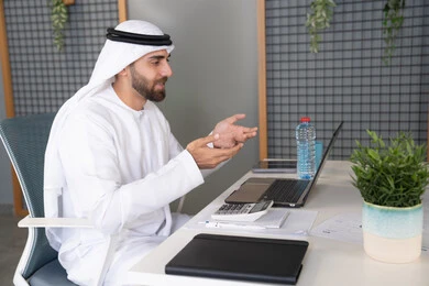 External business deals, online business meetings, business integration with technology, UAE companies, live broadcast and video calls, waving and greeting remotely, an Emirati Gulf Arab man wearing a ghutra and kandura using a laptop