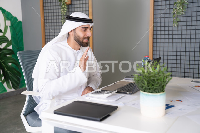 التلويح باليد والسلام عن بعد، رجل عربي خليجي اماراتي يرتدي الغترة والكندورة يقوم باستخدام جهاز الحاسوب المحمول, عقد صفقات عمل خارجية، اجتماعات عمل أونلاين، دمج العمل بالتقنية والتكنولوجيا، شركات الامارات العربية المتحدة، بث مباشر ومكالمات فيديو
