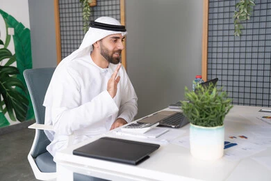 التلويح باليد والسلام عن بعد، رجل عربي خليجي اماراتي يرتدي الغترة والكندورة يقوم باستخدام جهاز الحاسوب المحمول, عقد صفقات عمل خارجية، اجتماعات عمل أونلاين، دمج العمل بالتقنية والتكنولوجيا، شركات الامارات العربية المتحدة، بث مباشر ومكالمات فيديو