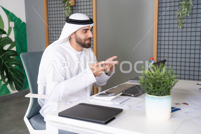 بث مباشر ومكالمات فيديو, التلويح باليد والسلام عن بعد، رجل عربي خليجي اماراتي يرتدي الغترة والكندورة يقوم باستخدام جهاز الحاسوب المحمول, عقد صفقات عمل خارجية، اجتماعات عمل أونلاين، دمج العمل بالتقنية والتكنولوجيا، شركات الامارات العربية المتحدة