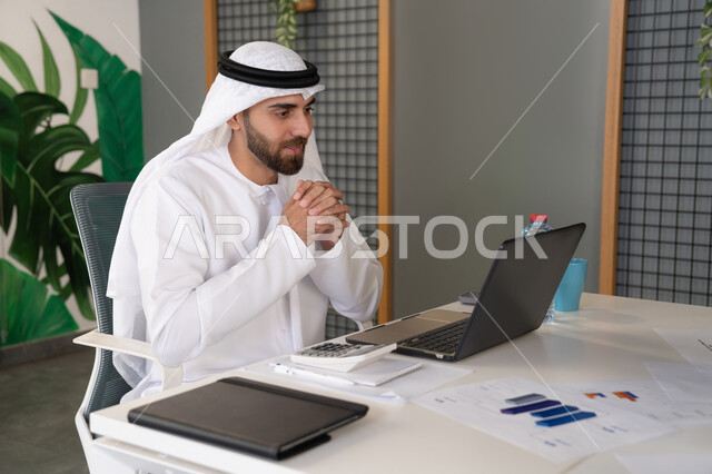 Integrating work and technology, UAE companies, live broadcasting and video calls, waving and greeting remotely, an Emirati Gulf Arab man wearing a ghutra and kandura using a laptop, conducting external business deals, online business meetings