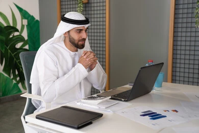Integrating work and technology, UAE companies, live broadcasting and video calls, waving and greeting remotely, an Emirati Gulf Arab man wearing a ghutra and kandura using a laptop, conducting external business deals, online business meetings