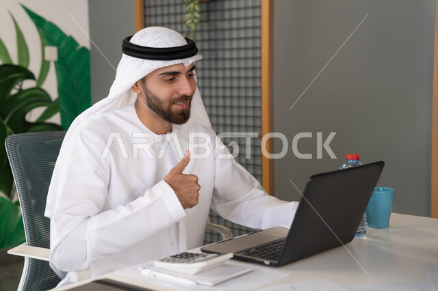 Integrating work and technology, UAE companies, live broadcasting and video calls, waving and greeting remotely, an Emirati Gulf Arab man wearing a ghutra and kandura using a laptop, conducting external business deals, online business meetings