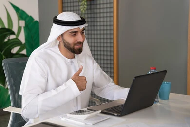 Integrating work and technology, UAE companies, live broadcasting and video calls, waving and greeting remotely, an Emirati Gulf Arab man wearing a ghutra and kandura using a laptop, conducting external business deals, online business meetings
