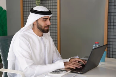 بث مباشر ومكالمات فيديو, التلويح باليد والسلام عن بعد، رجل عربي خليجي اماراتي يرتدي الغترة والكندورة يقوم باستخدام جهاز الحاسوب المحمول, عقد صفقات عمل خارجية، اجتماعات عمل أونلاين، دمج العمل بالتقنية والتكنولوجيا، شركات الامارات العربية المتحدة