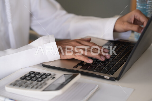 تحليل نتائج البيانات الاحصائية، صورة مقربة لرجل عربي خليجي اماراتي يرتدي الكندورة والغترة يعمل على الحاسب المحمول لانجاز المهام، تدوين الملاحظات على الورق، استخدام الالة الحاسبة لاجراء العمليات الحسابية، وظيفة مكتبية في شركات الامارات العربية المتحدة