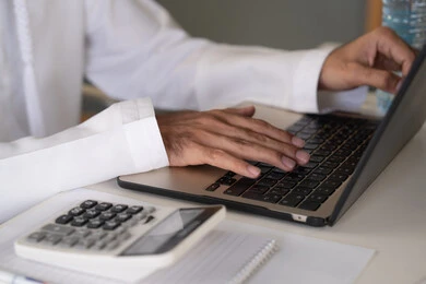 تحليل نتائج البيانات الاحصائية، صورة مقربة لرجل عربي خليجي اماراتي يرتدي الكندورة والغترة يعمل على الحاسب المحمول لانجاز المهام، تدوين الملاحظات على الورق، استخدام الالة الحاسبة لاجراء العمليات الحسابية، وظيفة مكتبية في شركات الامارات العربية المتحدة