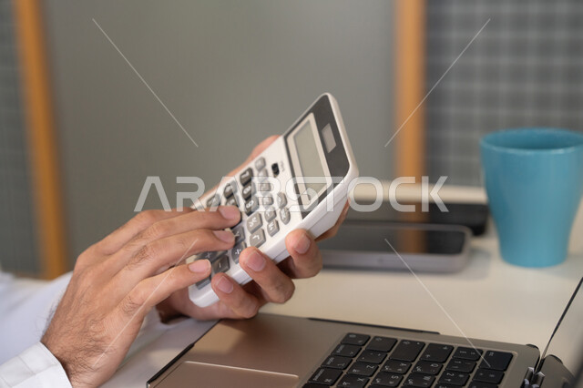 تحليل نتائج البيانات الاحصائية، صورة مقربة لرجل عربي خليجي اماراتي يرتدي الكندورة والغترة يعمل على الحاسب المحمول لانجاز المهام، تدوين الملاحظات على الورق، استخدام الالة الحاسبة لاجراء العمليات الحسابية، وظيفة مكتبية في شركات الامارات العربية المتحدة