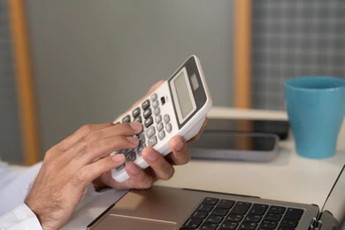 تحليل نتائج البيانات الاحصائية، صورة مقربة لرجل عربي خليجي اماراتي يرتدي الكندورة والغترة يعمل على الحاسب المحمول لانجاز المهام، تدوين الملاحظات على الورق، استخدام الالة الحاسبة لاجراء العمليات الحسابية، وظيفة مكتبية في شركات الامارات العربية المتحدة