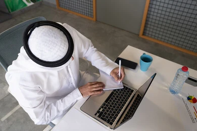 تدوين الملاحظات على الورق، استخدام الالة الحاسبة لاجراء العمليات الحسابية، وظيفة مكتبية في شركات الامارات العربية المتحدة، تحليل نتائج البيانات الاحصائية، صورة مقربة لرجل عربي خليجي اماراتي يرتدي الكندورة والغترة يعمل على الحاسب المحمول لانجاز المهام