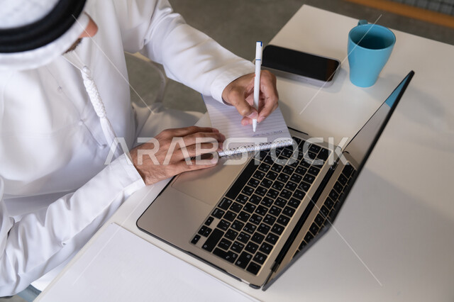 استخدام الالة الحاسبة لاجراء العمليات الحسابية، وظيفة مكتبية في شركات الامارات العربية المتحدة، تحليل نتائج البيانات الاحصائية، صورة مقربة لرجل عربي خليجي اماراتي يرتدي الكندورة والغترة يعمل على الحاسب المحمول لانجاز المهام، تدوين الملاحظات على الورق