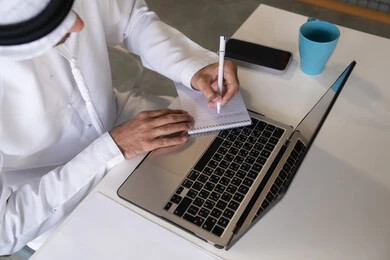 استخدام الالة الحاسبة لاجراء العمليات الحسابية، وظيفة مكتبية في شركات الامارات العربية المتحدة، تحليل نتائج البيانات الاحصائية، صورة مقربة لرجل عربي خليجي اماراتي يرتدي الكندورة والغترة يعمل على الحاسب المحمول لانجاز المهام، تدوين الملاحظات على الورق