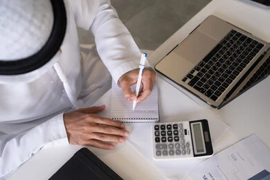 تدوين الملاحظات على الورق، استخدام الالة الحاسبة لاجراء العمليات الحسابية، وظيفة مكتبية في شركات الامارات العربية المتحدة، تحليل نتائج البيانات الاحصائية، صورة مقربة لرجل عربي خليجي اماراتي يرتدي الكندورة والغترة يعمل على الحاسب المحمول لانجاز المهام