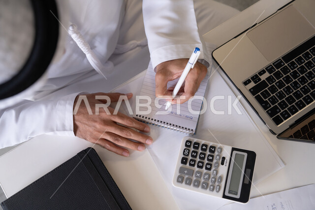 تدوين الملاحظات على الورق، استخدام الالة الحاسبة لاجراء العمليات الحسابية، وظيفة مكتبية في شركات الامارات العربية المتحدة، تحليل نتائج البيانات الاحصائية، صورة مقربة لرجل عربي خليجي اماراتي يرتدي الكندورة والغترة يعمل على الحاسب المحمول لانجاز المهام