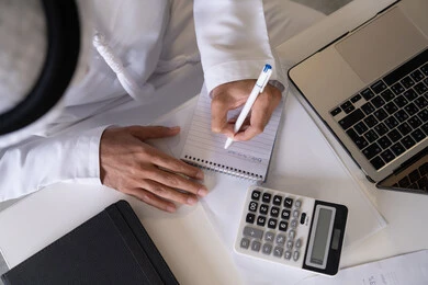 Taking notes on paper, using a calculator to perform calculations, office job in UAE companies, analyzing statistical data results, close-up of an Emirati Gulf Arab man wearing a kandura and ghutra working on a laptop to complete tasks