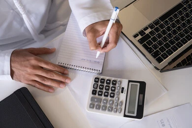 استخدام الالة الحاسبة لاجراء العمليات الحسابية، وظيفة مكتبية في شركات الامارات العربية المتحدة، تحليل نتائج البيانات الاحصائية، صورة مقربة لرجل عربي خليجي اماراتي يرتدي الكندورة والغترة يعمل على الحاسب المحمول لانجاز المهام، تدوين الملاحظات على الورق