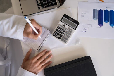 استخدام الالة الحاسبة لاجراء العمليات الحسابية، وظيفة مكتبية في شركات الامارات العربية المتحدة، تحليل نتائج البيانات الاحصائية، صورة مقربة لرجل عربي خليجي اماراتي يرتدي الكندورة والغترة يعمل على الحاسب المحمول لانجاز المهام، تدوين الملاحظات على الورق