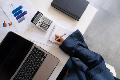 كتابة وتدوين الملاحظات على الورق، استخدام الالة الحاسبة لاجراء العمليات الحسابية، وظيفة مكتبية في شركات الامارات العربية المتحدة، تحليل نتائج البيانات الاحصائية، صورة مقربة لإمرأة عربية خليجية اماراتية تعمل على الحاسب المحمول لانجاز المهام