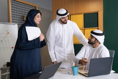 دراسة و تطوير المشاريع الجديدة في شركات الامارات العربية المتحدة، استخدام جهاز تقني حديث، جمع البيانات وحل المشكلات، اجتماع ومناقشة عمل في مقر الشركة، رجل أعمال عربي خليجي اماراتي يرتدي الكندورة والغترة يقوم بالعمل على الحاسب المحمول مع زملاءه داخل المكتب