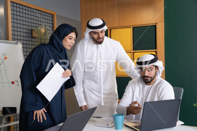 Developing and studying new projects in UAE companies, using modern technical equipment, collecting data and solving problems, meeting and discussing work at the company headquarters, an Emirati Gulf Arab businessman wearing a kandura and ghutra working on a laptop with his colleagues inside the office