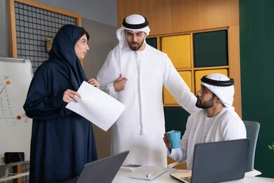 Achieving company goals, meeting and discussing work at company headquarters, an Emirati Gulf Arab businessman wearing a kandura and ghutra working on a laptop with his colleagues inside the office, studying and developing new projects in UAE companies, using a modern technical device