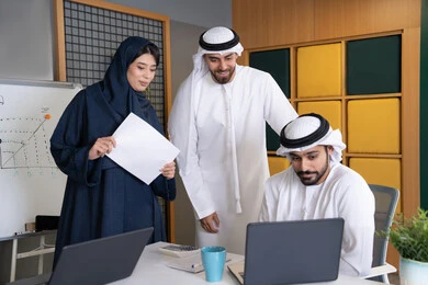 حل المشكلات وتحليل البيانات، اجتماع ومناقشة عمل في مقر الشركة، رجل أعمال عربي خليجي اماراتي يرتدي الكندورة والغترة يقوم بالعمل على الحاسب المحمول مع زملاءه داخل المكتب، دراسة و تطوير المشاريع الجديدة في شركات الامارات العربية المتحدة، استخدام جهاز تقني حديث