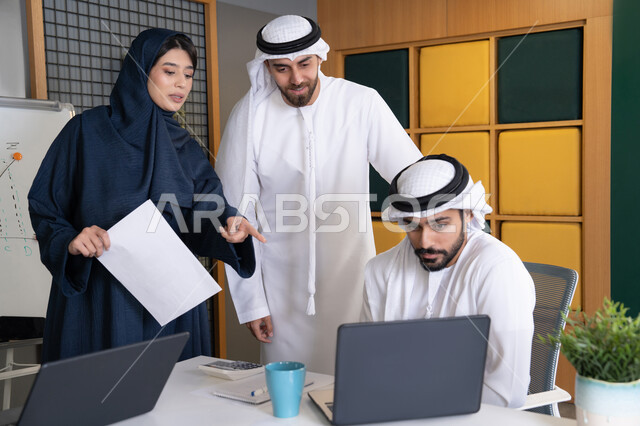 تحقيق اهداف الشركة، اجتماع ومناقشة عمل في مقر الشركة، رجل أعمال عربي خليجي اماراتي يرتدي الكندورة والغترة يقوم بالعمل على الحاسب المحمول مع زملاءه داخل المكتب، دراسة و تطوير المشاريع الجديدة في شركات الامارات العربية المتحدة، استخدام جهاز تقني حديث