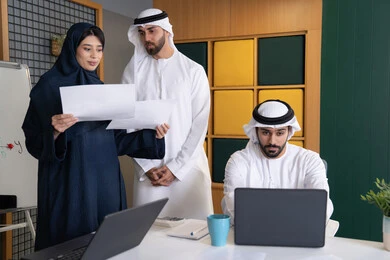 Studying and developing new projects in UAE companies, using modern technical equipment, collecting data and solving problems, meeting and discussing work at the company headquarters, an Emirati Gulf Arab businessman wearing a kandura and ghutra working on a laptop with his colleagues inside the office