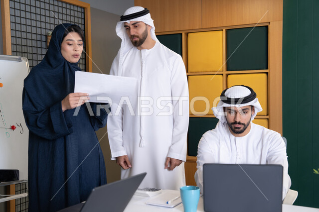 حل المشكلات وتحليل البيانات، اجتماع ومناقشة عمل في مقر الشركة، رجل أعمال عربي خليجي اماراتي يرتدي الكندورة والغترة يقوم بالعمل على الحاسب المحمول مع زملاءه داخل المكتب، دراسة و تطوير المشاريع الجديدة في شركات الامارات العربية المتحدة، استخدام جهاز تقني حديث