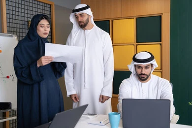 حل المشكلات وتحليل البيانات، اجتماع ومناقشة عمل في مقر الشركة، رجل أعمال عربي خليجي اماراتي يرتدي الكندورة والغترة يقوم بالعمل على الحاسب المحمول مع زملاءه داخل المكتب، دراسة و تطوير المشاريع الجديدة في شركات الامارات العربية المتحدة، استخدام جهاز تقني حديث