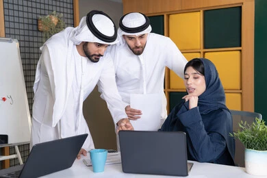 Work environment in UAE companies, using modern technology, achieving company goals, meeting and discussing work at company headquarters, an Emirati Gulf Arab businesswoman wearing a hijab and abaya working on a laptop with her colleagues inside the office, studying and developing new projects