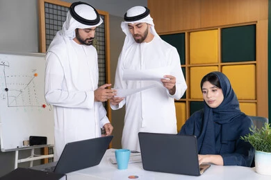 بيئة العمل في شركات الامارات العربية المتحدة، استخدام جهاز تقني حديث، تحقيق اهداف الشركة، اجتماع ومناقشة عمل في مقر الشركة، سيدة أعمال عربية خليجية اماراتية ترتدي الحجاب والعباءة تعمل على الحاسب المحمول مع زملائها داخل المكتب، دراسة و تطوير المشاريع الجديدة