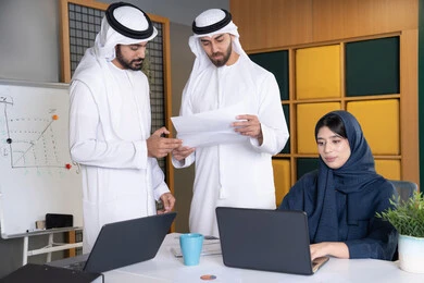 تطوير ودراسة المشاريع الجديدة في شركات الامارات العربية المتحدة، استخدام جهاز تقني حديث، جمع البيانات وحل المشكلات، اجتماع ومناقشة عمل في مقر الشركة، رجل أعمال عربي خليجي اماراتي يرتدي الكندورة والغترة يقوم بالعمل على الحاسب المحمول مع زملاءه داخل المكتب
