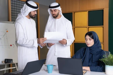 Work environment in UAE companies, using modern technology, achieving company goals, meeting and discussing work at company headquarters, an Emirati Gulf Arab businesswoman wearing a hijab and abaya working on a laptop with her colleagues inside the office, studying and developing new projects