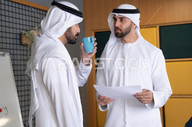 عرض مزايا الصفقات الجديدة، مفهوم الإدارة المتعاونة، موظفان عربيان خليجيان اماراتيان يرتديان الكندورة والغترة يناقشان أمور العمل باستخدام الأوراق والبيانات، الوحدة والتفاعل الجماعي لتحقيق أهداف الشركة وحل المشكلات في شركات الامارات العربية المتحدة
