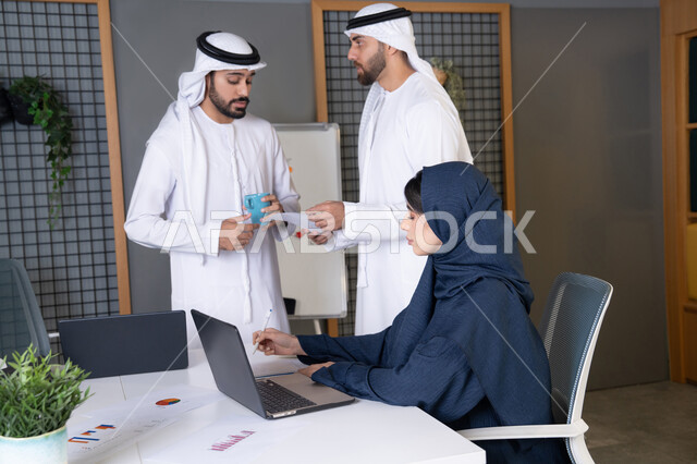دراسة و تطوير المشاريع الجديدة، بيئة العمل في شركات الامارات العربية المتحدة، استخدام جهاز تقني حديث، تحقيق اهداف الشركة، اجتماع ومناقشة عمل في مقر الشركة، سيدة أعمال عربية خليجية اماراتية ترتدي الحجاب والعباءة تعمل على الحاسب المحمول مع زملائها داخل المكتب